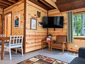 A wooden cabin interior with a television, a dining table, chairs, and a window with a view of trees.