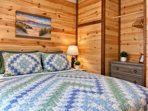 A wooden-paneled room with a bed, a nightstand, a lamp, and a painting of a beach scene.