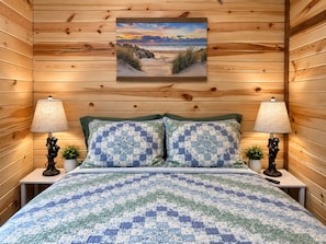 A room with wooden panel walls, a bed with a blue and white quilt, two bedside lamps, and a framed picture on the wall.