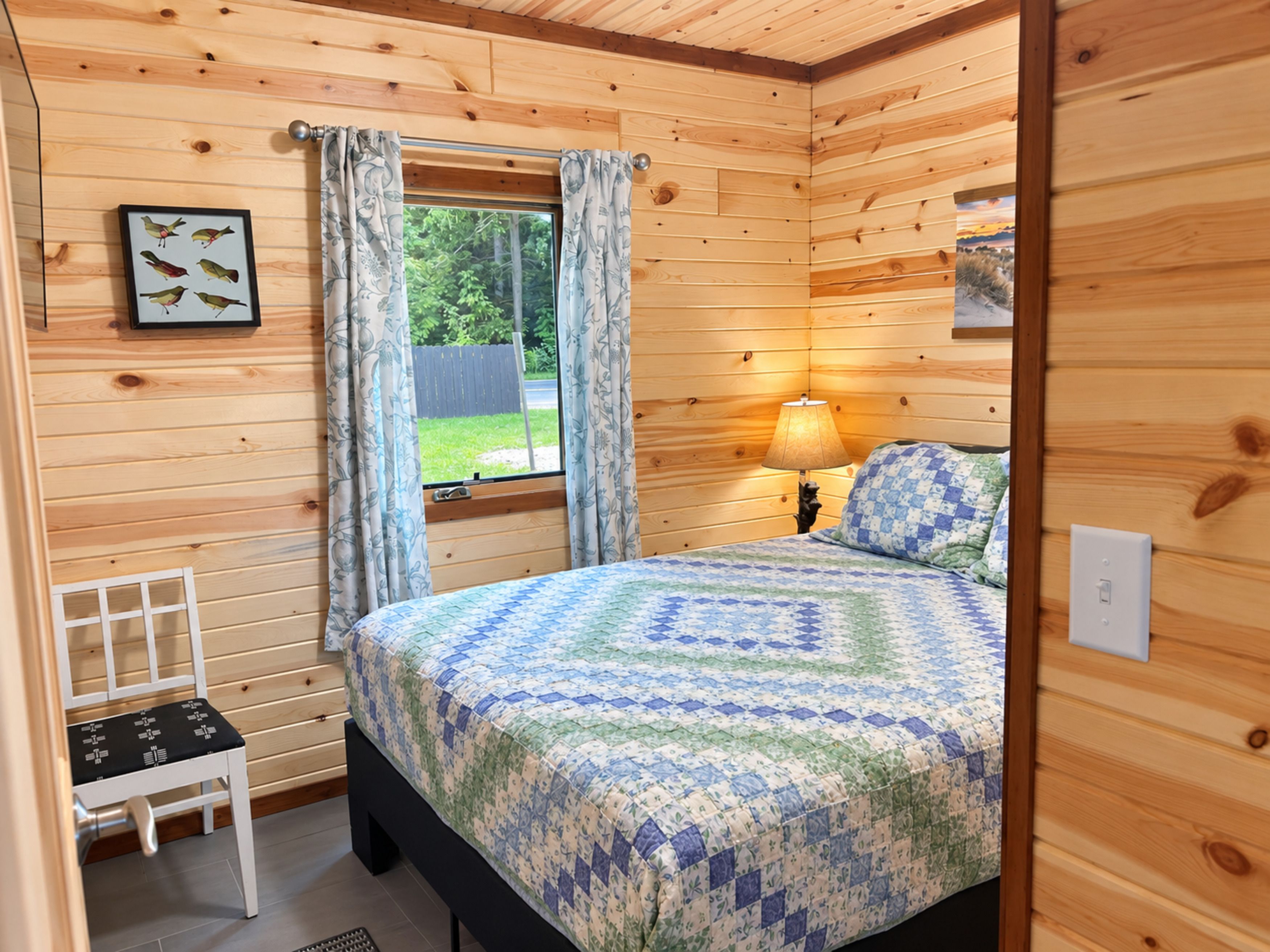 A wooden-paneled room with a bed, a window with curtains, a bedside lamp, and a framed picture on the wall.