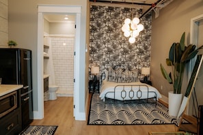 A bedroom with a patterned wallpaper, a bed with a headboard, bedside lamps, a large potted plant, and a hanging light fixture.