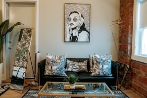 Un salon moderne avec un canapé noir, des coussins à motifs, une table basse en verre, une grande œuvre d’art encadrée et une plante en pot.