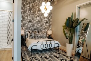 Une chambre avec un papier peint à motifs, un lit avec un cadre en métal, deux lampes de chevet, une grande plante en pot et un miroir appuyé contre le mur.