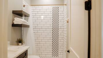 Un baño con ducha de azulejos blancos, inodoro, lavamanos con encimera de mármol y estante con toallas.
