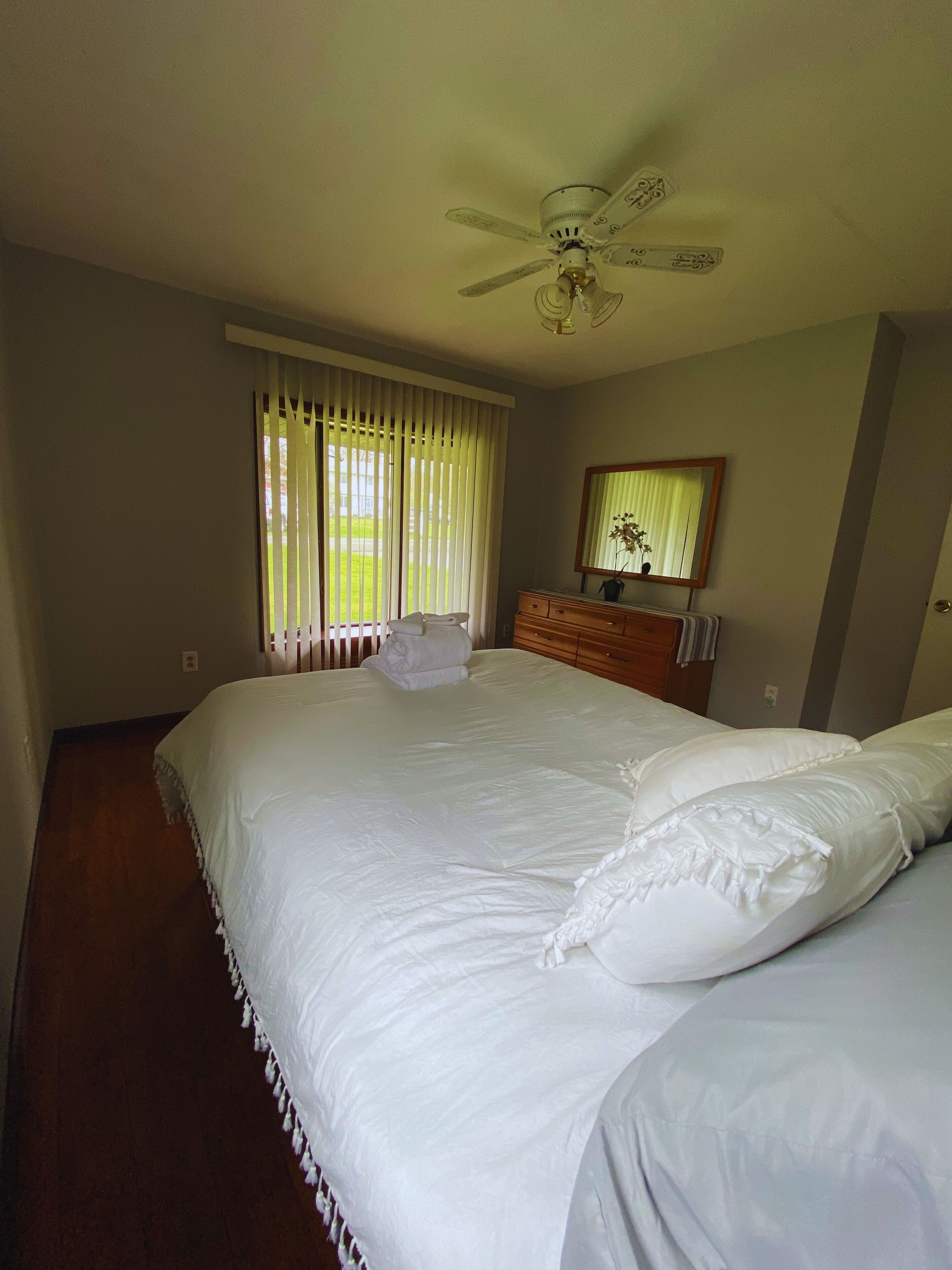 Una camera da letto con un letto grande, un ventilatore a soffitto, una finestra con tende, un comò e uno specchio.