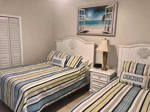 A bedroom with two beds, each with striped bedding and a pillow labeled "BEACH". There is a nightstand with a lamp and a small table with a shell decoration. A painting of an open window with a beach scene is on the wall. - Splash Pool- Key Colony Villa - KCB#4 (Key Colony Beach)
