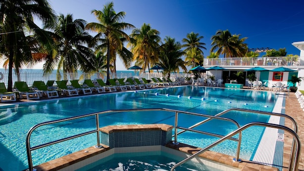 A swimming pool with lounge chairs and palm trees, surrounded by a fence and a building with a balcony. - Splash Pool- Key Colony Villa - KCB#4 (Key Colony Beach)