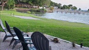 Espace extérieur aménagé avec des chaises et des tables sur une surface de gravier, à proximité d’un plan d’eau, avec une jetée et de la verdure en arrière-plan.
