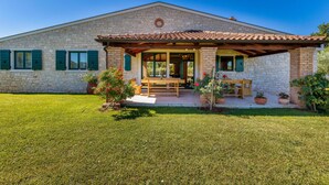 Un bâtiment en pierre avec un toit en tuiles, un patio couvert avec une table et des chaises, et une pelouse bien entretenue.