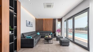 Una sala de estar moderna con un sofá seccional, una mesa de centro con frutas y vasos, y vista a una piscina a través de grandes ventanales.