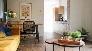 Un soggiorno moderno con un divano giallo, poltrone in vimini, un tavolo da pranzo e un angolo cottura con pensili in legno e una macchina per il caffè.