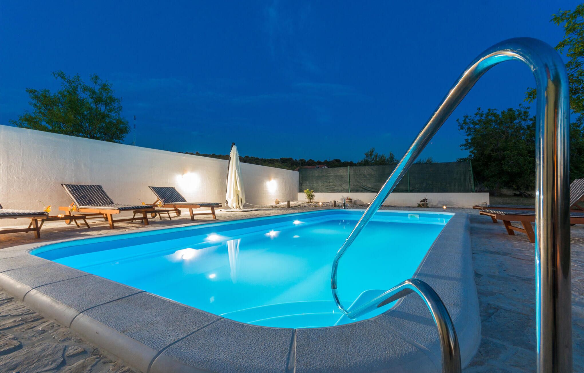 Una piscina con una escalera metálica, sillas de descanso y una pared blanca al fondo.