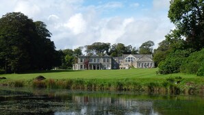 A large, multi-story building with a symmetrical facade, situated on a lush green landscape with a pond in the foreground. - Exceptional 3-bedroom lodge on spacious country estate. Free gym, pool & more. (Praze-an-Beeble)