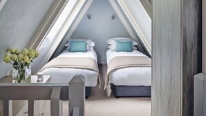 A bedroom with two beds, a small table with a vase of flowers, and a view of the sky through a triangular window. - Exceptional 3-bedroom lodge on spacious country estate. Free gym, pool & more. (Praze-an-Beeble)