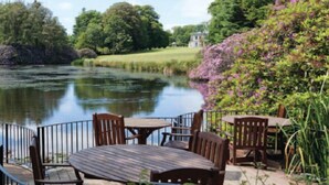A riverside patio with wooden tables and chairs, surrounded by lush greenery and a calm river. - Exceptional 3-bedroom lodge on spacious country estate. Free gym, pool & more. (Praze-an-Beeble)