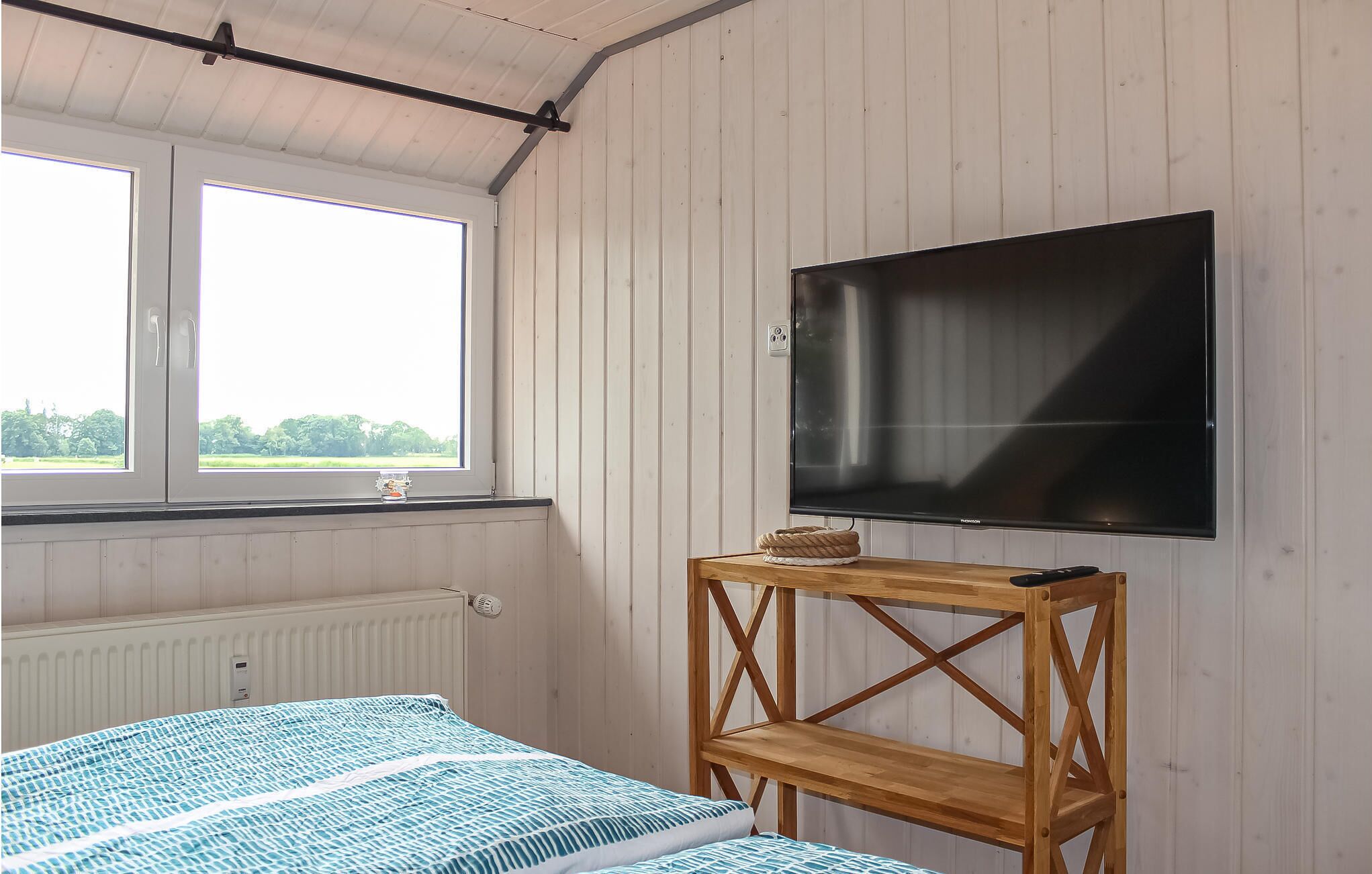 A room with a bed, a wooden shelf, a TV, and a window with a view of greenery.
