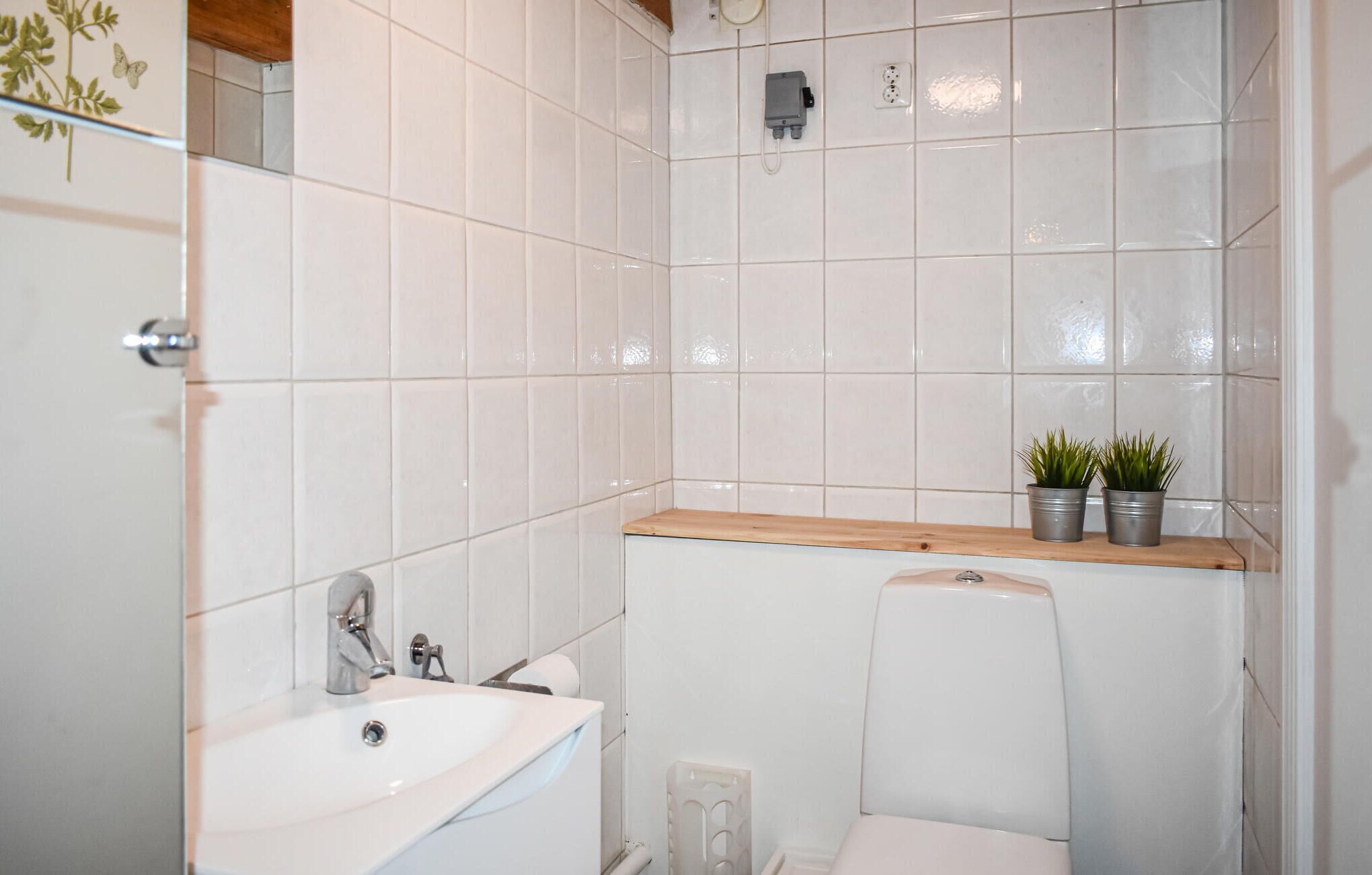 A small bathroom with white tiles, a sink, a toilet, and a shelf with two potted plants.