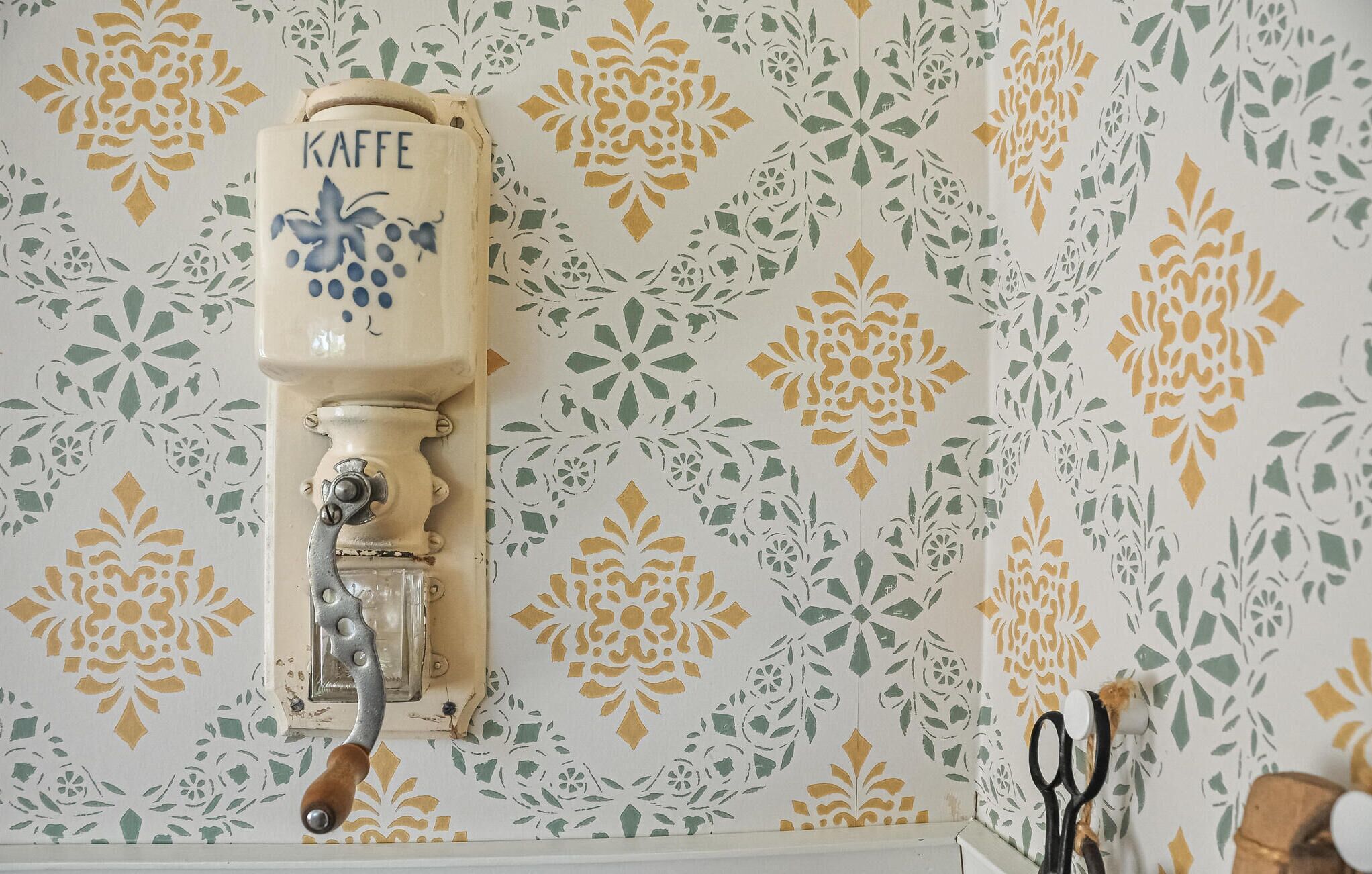 A vintage coffee grinder with the word "KAFFE" on it, mounted on a wall with a floral patterned wallpaper.