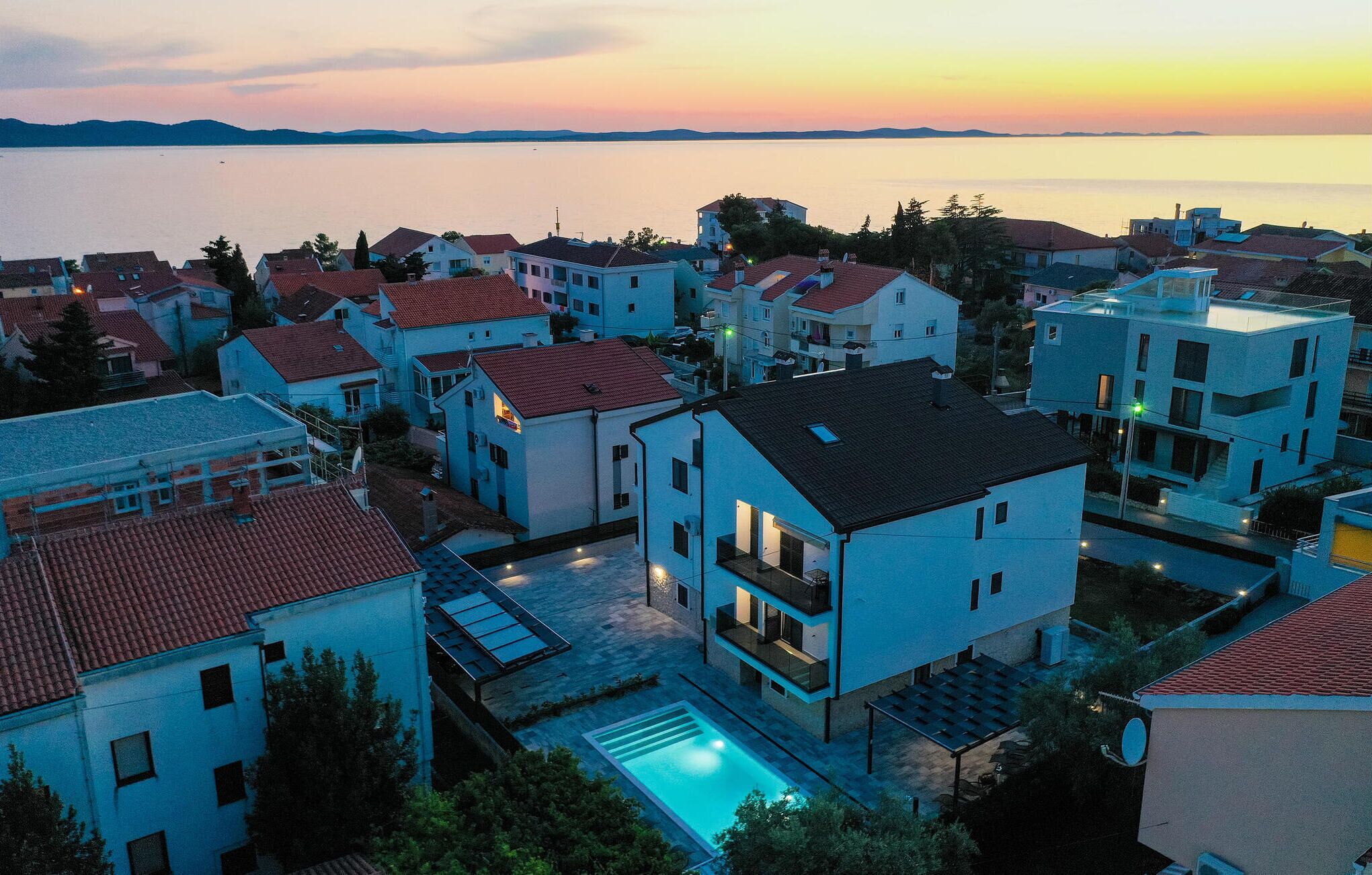 Zona residencial con casas modernas, una piscina y vistas al mar al atardecer.