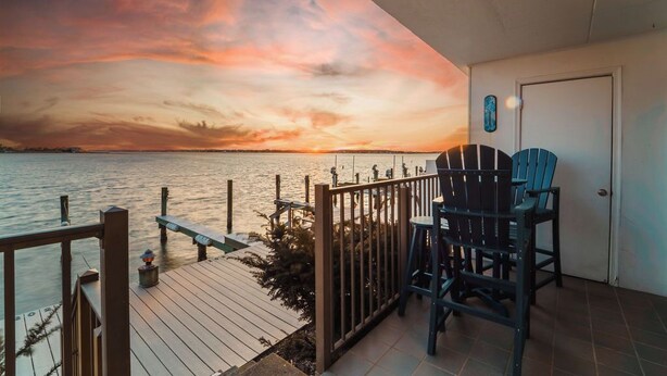 Une terrasse en bois offrant une vue sur un coucher de soleil sur l’eau, deux fauteuils Adirondack et une porte agrémentée d’une petite plante en pot.