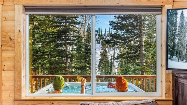 Une cabane dotée d’une grande fenêtre donnant sur une forêt. À l’intérieur, on trouve un jacuzzi et une terrasse en bois.