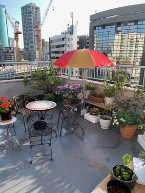 A rooftop terrace with a table, chairs, potted plants, and a red umbrella.
