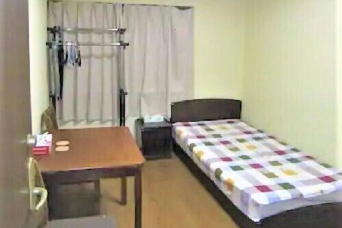 A small, well-lit room with a single bed, a desk, and a chair. There is a window with curtains, a radiator, and a ceiling light.