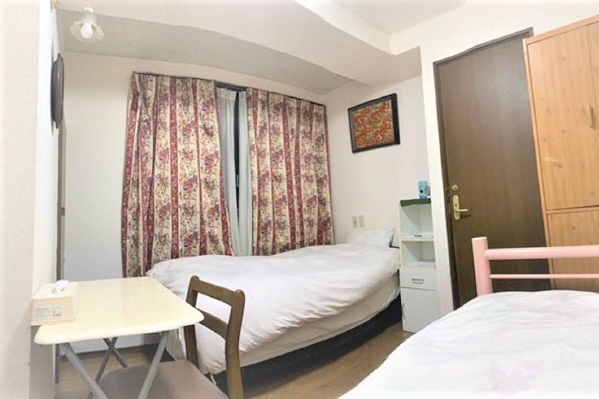 A small bedroom with two single beds, a wooden wardrobe, a desk with a chair, and a window with floral curtains.