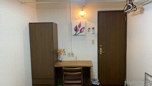 Una habitación pequeña con un escritorio de madera, una silla, un florero con flores, una lámpara en la pared y una puerta.