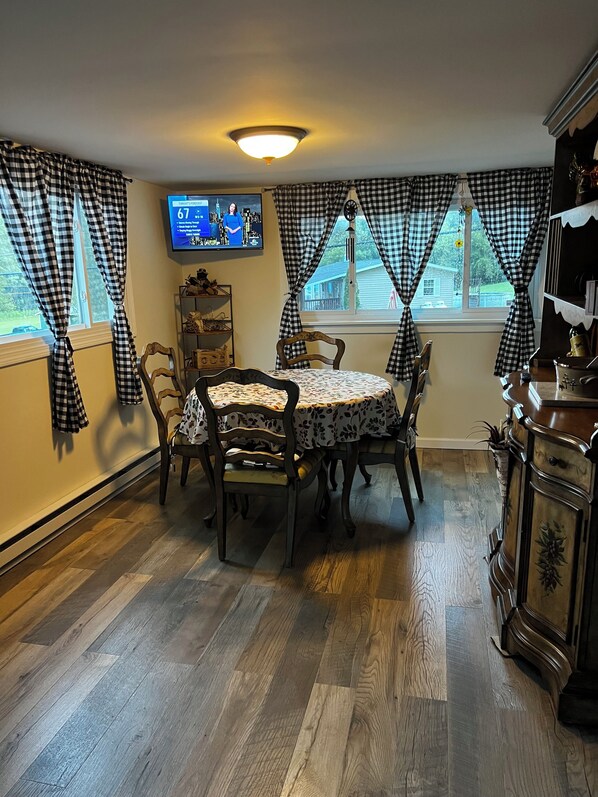 A dining area with a round table, four chairs, a television mounted on the wall, and checkered curtains. - Cozy County Home (Richfield Springs)