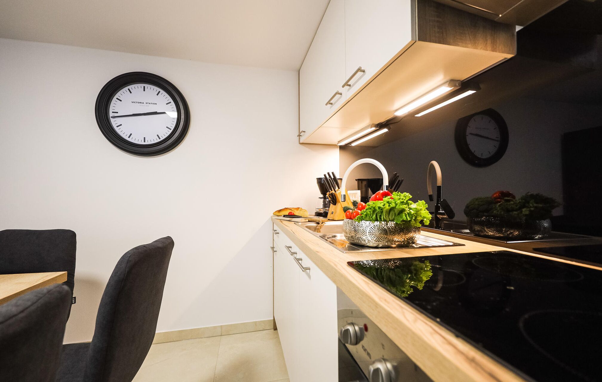 A modern kitchen with a built-in stove, a sink, and a countertop with a plant. There is a clock on the wall and a chair in the corner.