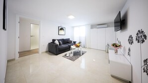 Een moderne woonkamer met een grijze bank, een salontafel met fruit, en een flatscreen televisie aan de muur.