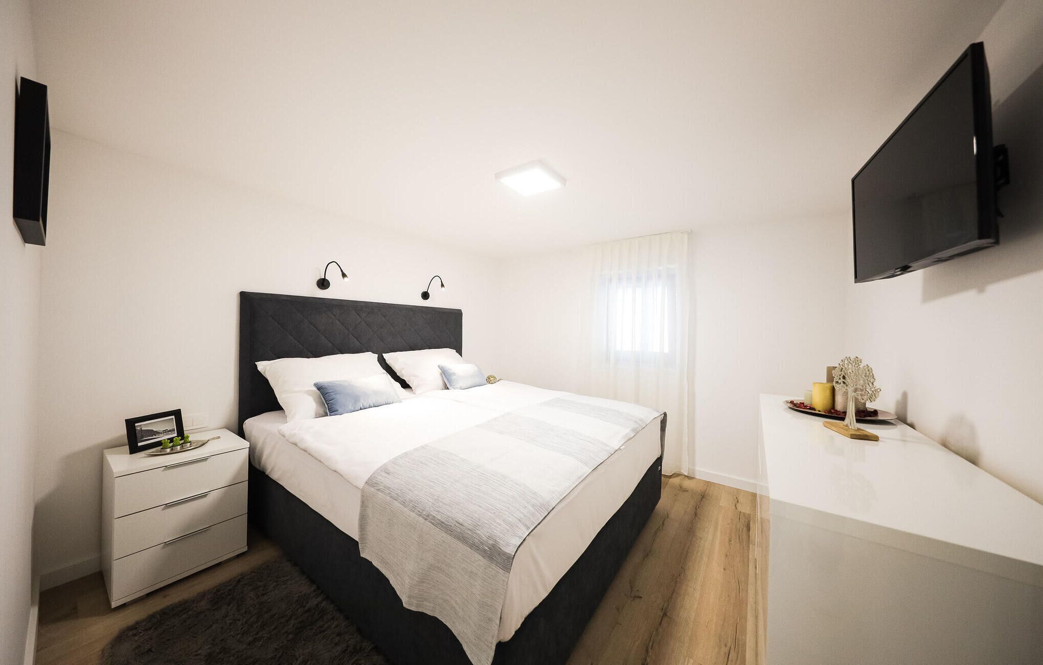 Quarto moderno com cama, mesas de cabeceira, televisão e uma pequena mesa com objetos decorativos.