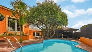 A swimming pool with a wooden deck and metal ladder, surrounded by a brick building with large windows and a covered patio area.