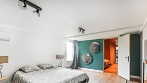 A bedroom with a bed, a bedside table, a lamp, a mirror, and a door leading to another room.