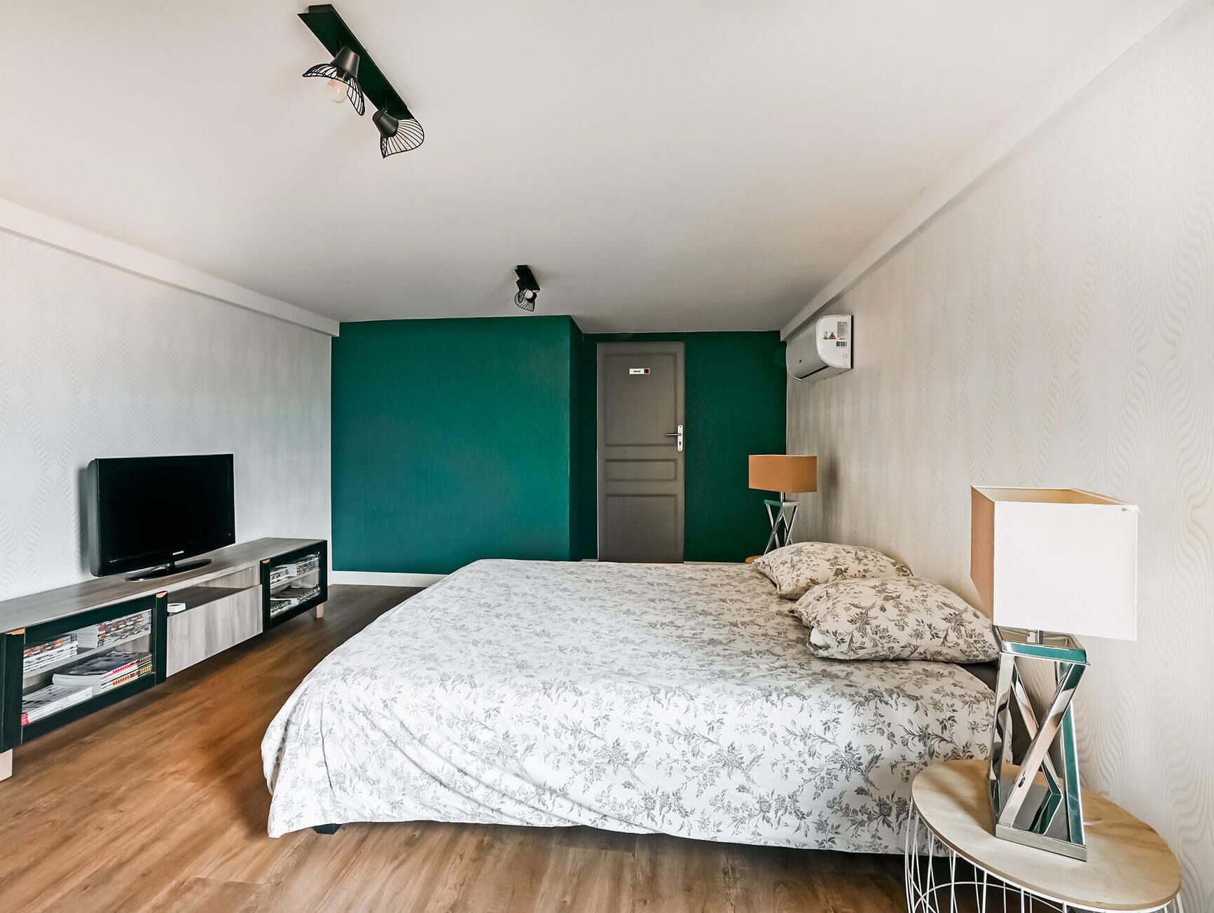 A hotel room with a bed, a TV, a bookshelf, a door, and a wall-mounted air conditioner.