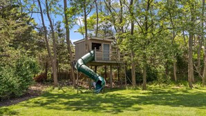Une cabane dans les arbres avec un toboggan, entourée d’une zone herbeuse et d’arbres.