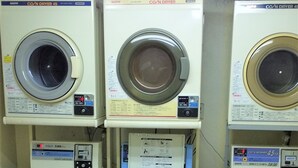 A laundry facility with three stacked washers and dryers, a control panel, and a chair. - Urban Times Hiroshima (Hiroshima)