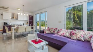 Een moderne woonkamer met een paarse bank, een eetgedeelte met een witte tafel, en een keuken met witte kasten en een roestvrijstalen afzuigkap.