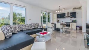 Een moderne woonkamer met een zwart-wit bank, een eethoek met witte stoelen, een keuken met zwarte kastjes en grote ramen met uitzicht op groen.