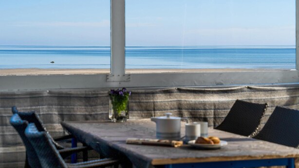 Une vue sur la mer depuis une fenêtre, avec une table dressée pour deux personnes, un coussin et un vase rempli de fleurs.