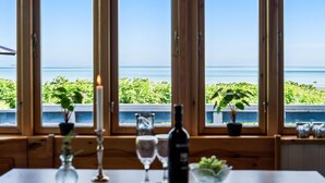Une table à manger, agrémentée d’une bougie, de verres à vin et d’une bouteille, est placée devant de grandes fenêtres qui offrent une vue imprenable sur un paysage verdoyant et luxuriant et sur la mer.