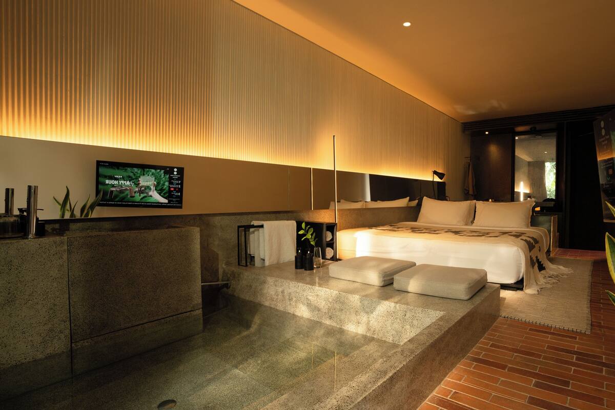Una habitación de hotel moderna con una cama grande, un área de descanso con una mesita, un televisor montado en la pared y un ventanal amplio.