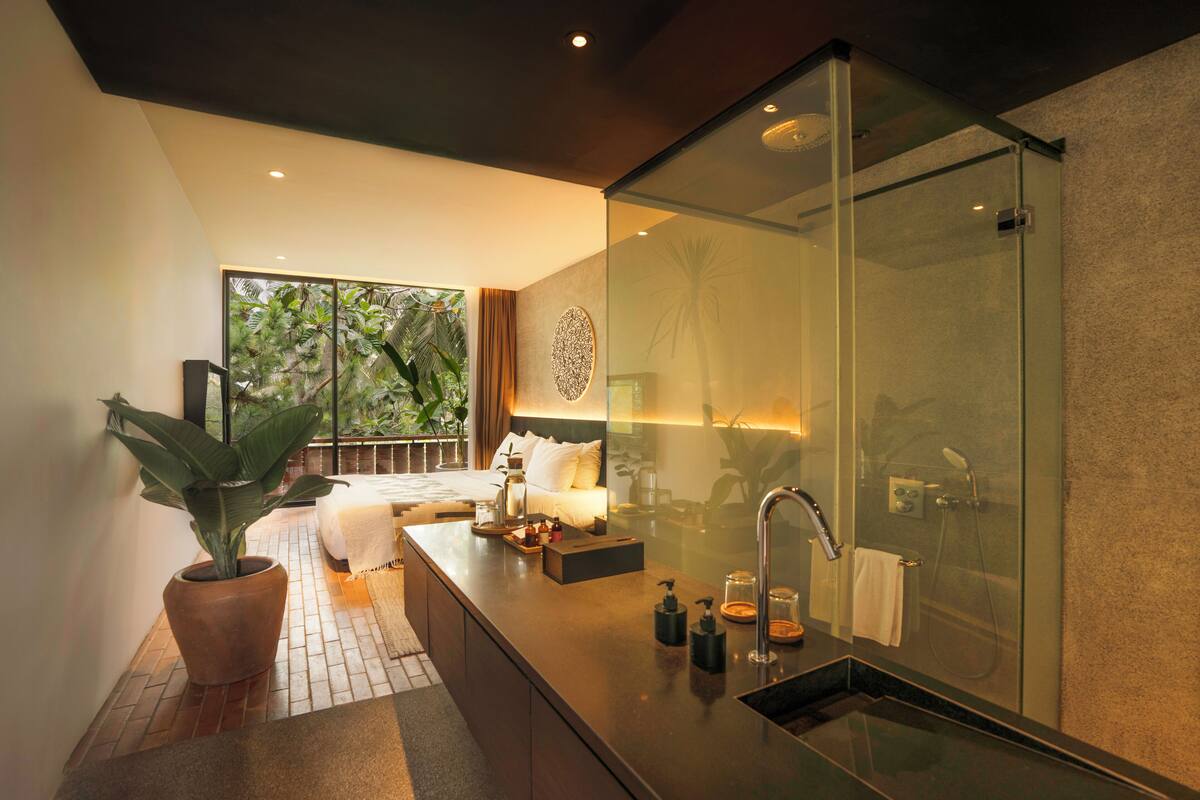 Un baño moderno con cabina de ducha de vidrio, una planta grande en maceta y una mesita de noche con lámpara.