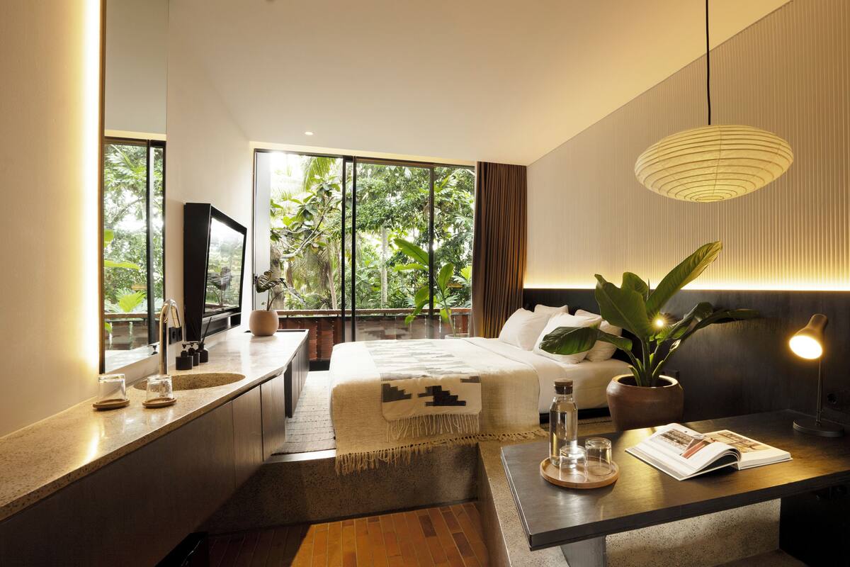 Un dormitorio moderno con una ventana grande, una cama, una mesita de noche con una revista, una planta y una lámpara colgante.