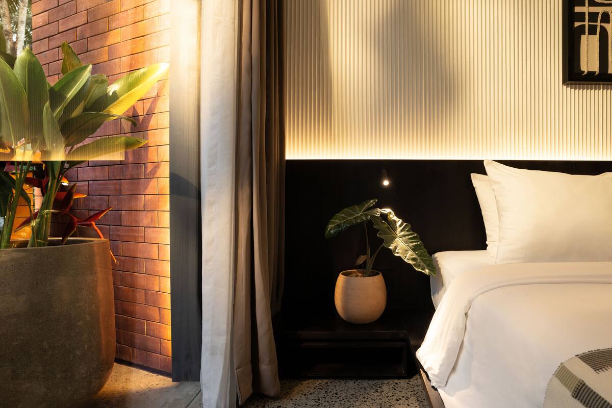 Habitación de hotel con pared de ladrillo, una planta en maceta, cama con ropa blanca y una obra enmarcada en la pared.