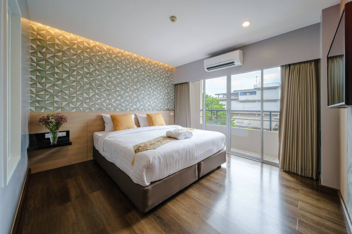 Quarto de hotel moderno com uma cama grande, parede com padrão geométrico e vista para a cidade através de amplas janelas.