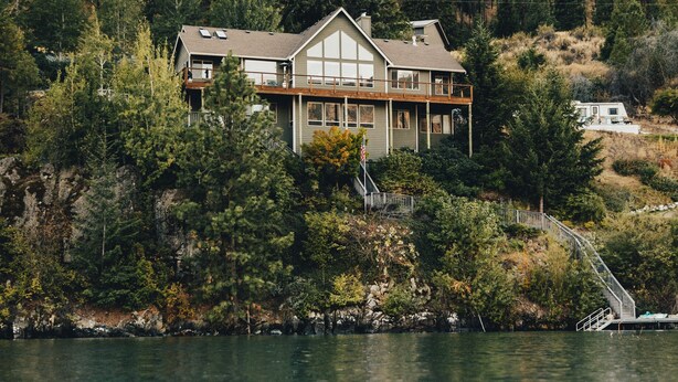 A large, multi-story house with a balcony, situated on a lakeside with a rocky backdrop.