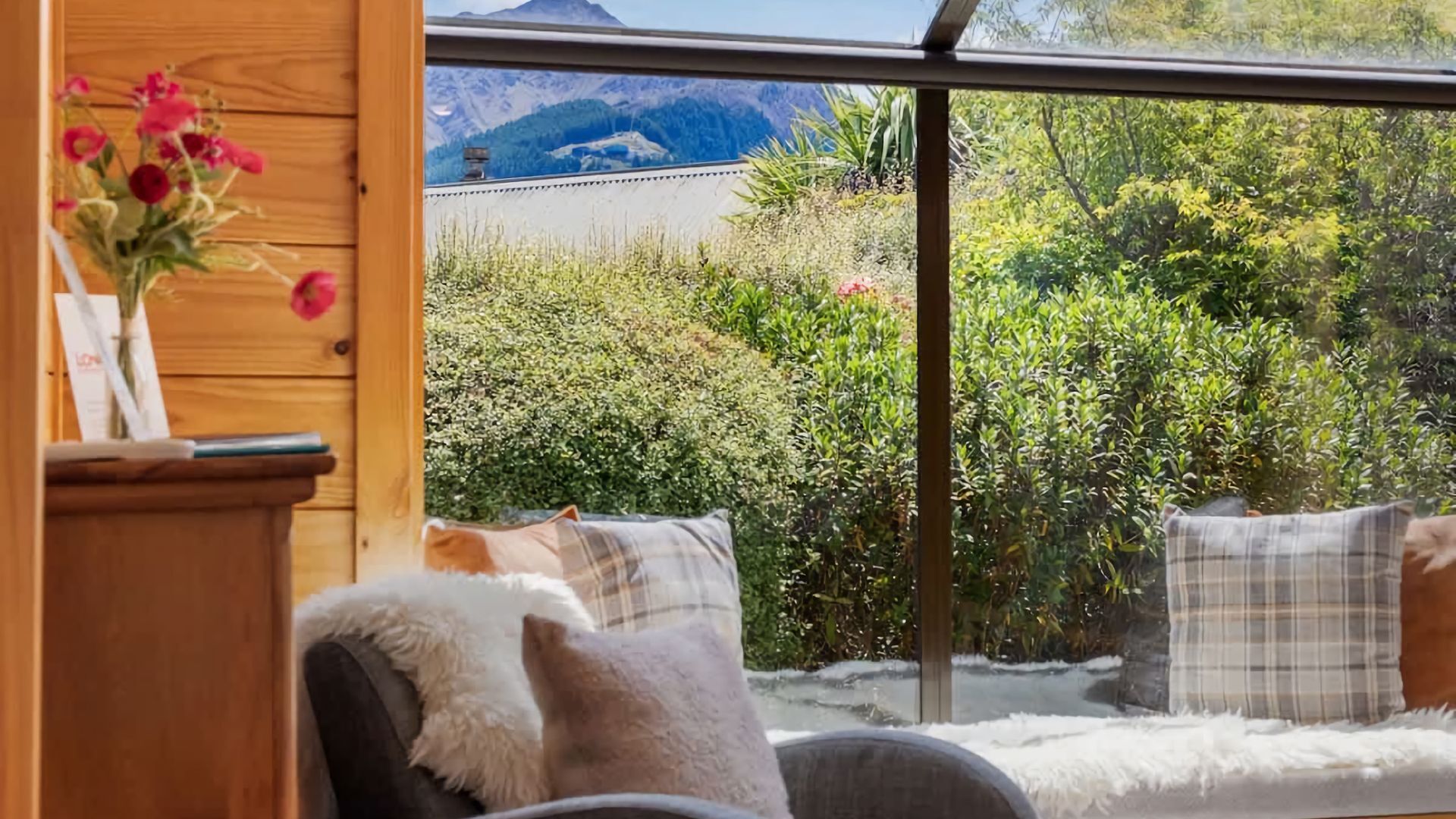 Habitación con un ventanal grande que ofrece vistas a un paisaje montañoso. En el interior, hay un armario de madera con un jarrón de flores y una cama con ropa de cama escocesa.