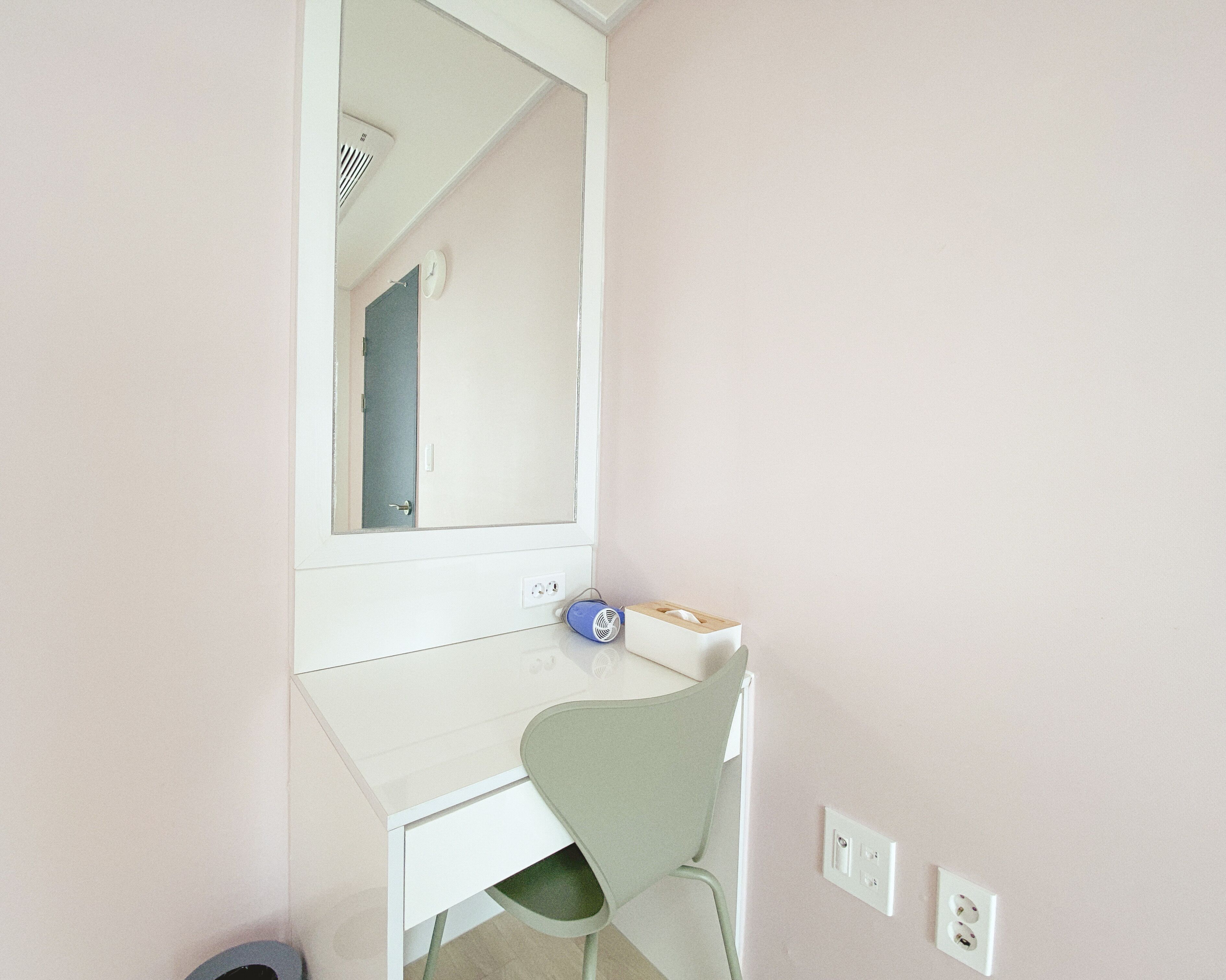 Una habitación pequeña y privada con un escritorio blanco, una silla verde, un espejo y un reloj en la pared.